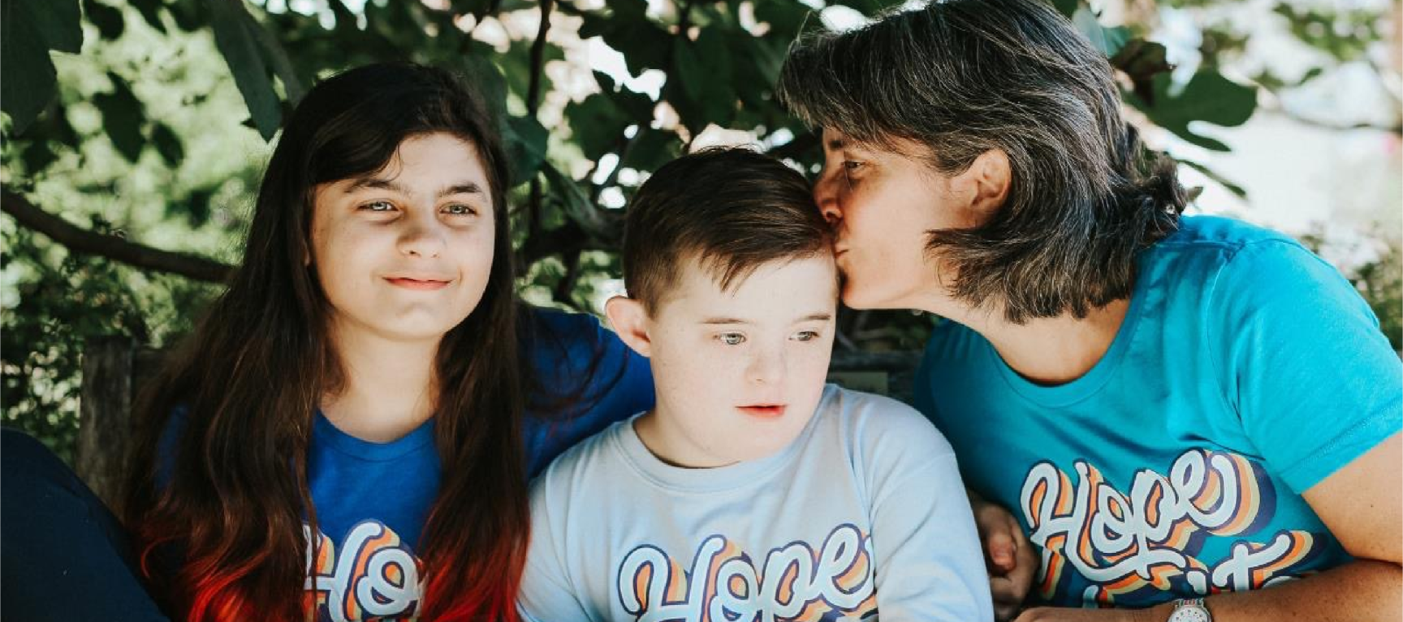 A mother and her two children sit together under a tree canopy wearing blue t-shirts with the word "Hope" on them. The mother is kissing the younger child, who has Down's Syndrome.